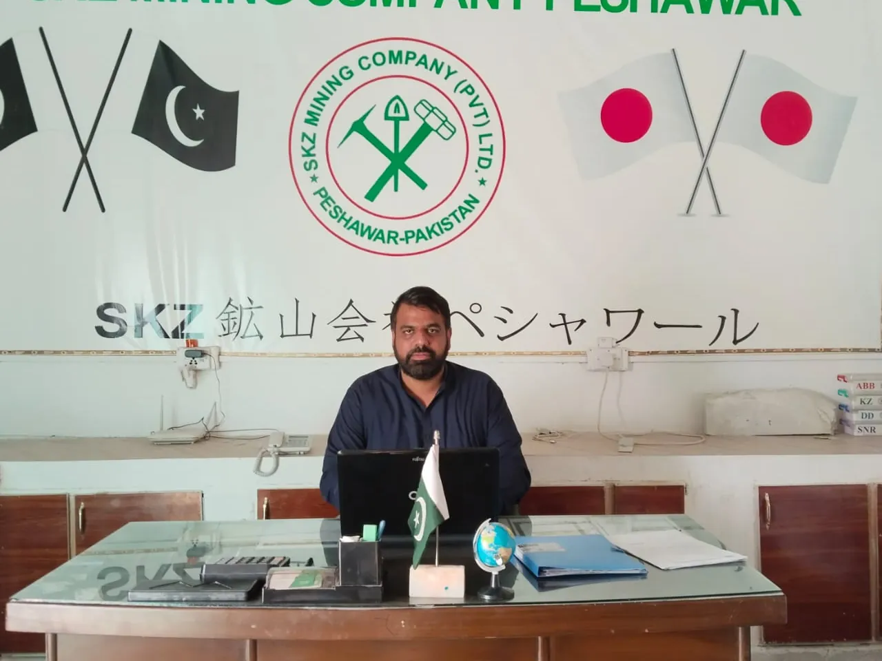 Manager seated at his desk in the SKZ Mining Company Peshawar office beneath the company sign.