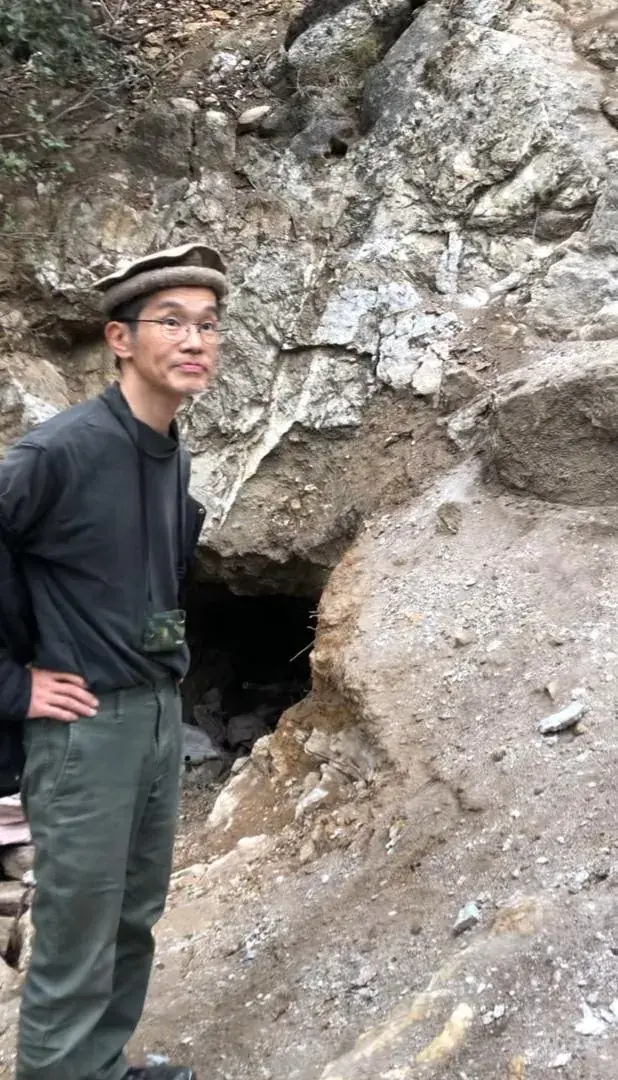 Japanese technical expert standing beside a small adit entrance in a rock face.