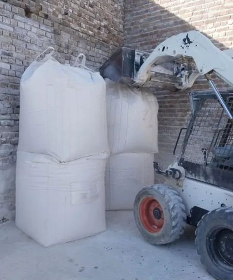 Skid-steer loader lifting white jumbo bags beside a brick wall.