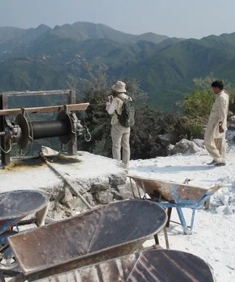 Hilltop mine site with a winch, wheelbarrows and visitors, green hills behind.