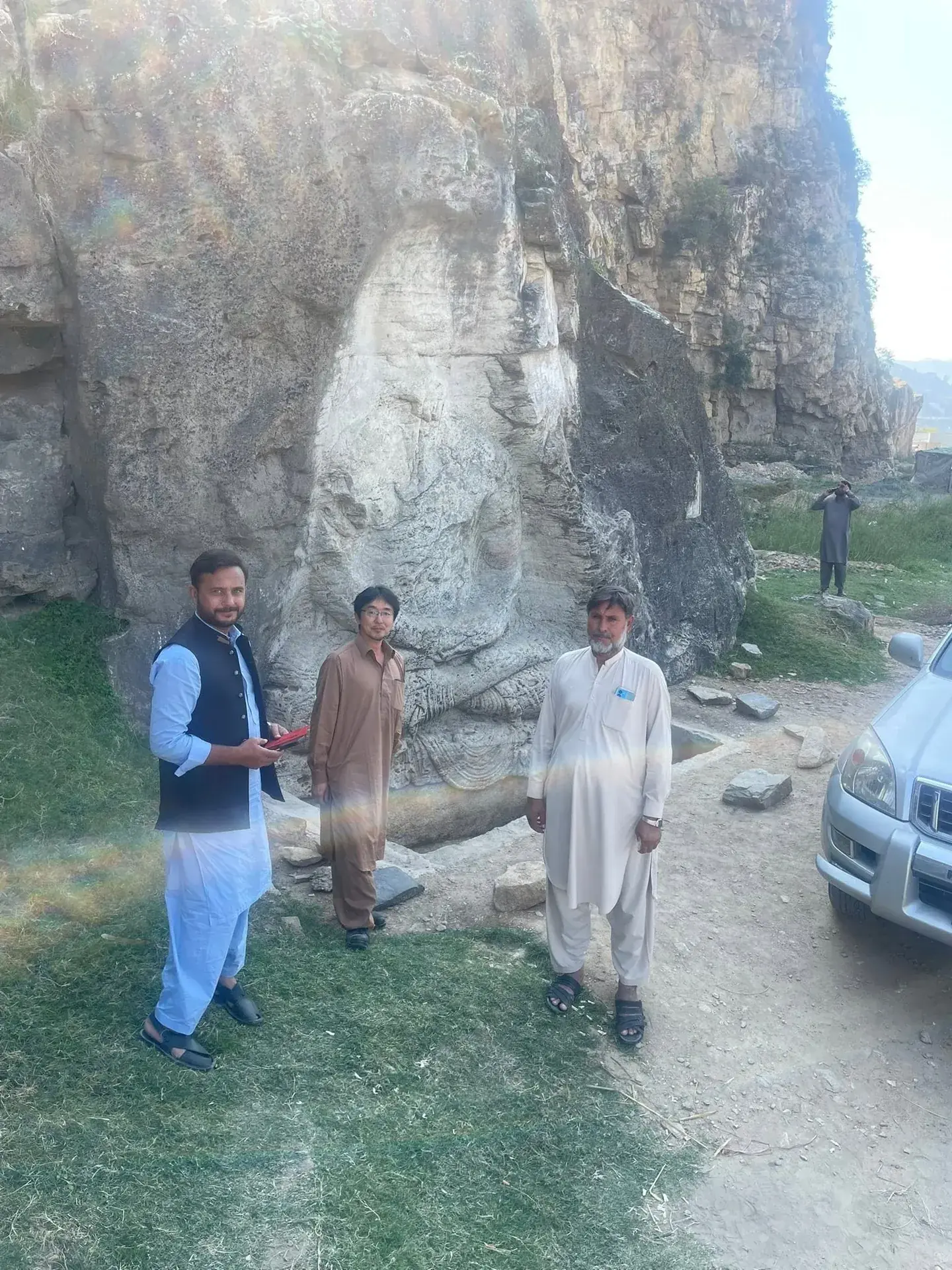 Three men standing beside a white rock outcrop on a hillside road with a parked vehicle.