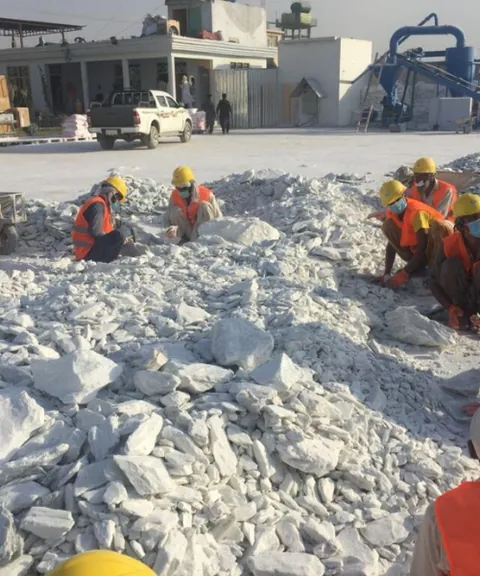 Workers in hard hats hand-sorting a pile of white talc lumps near a plant building.