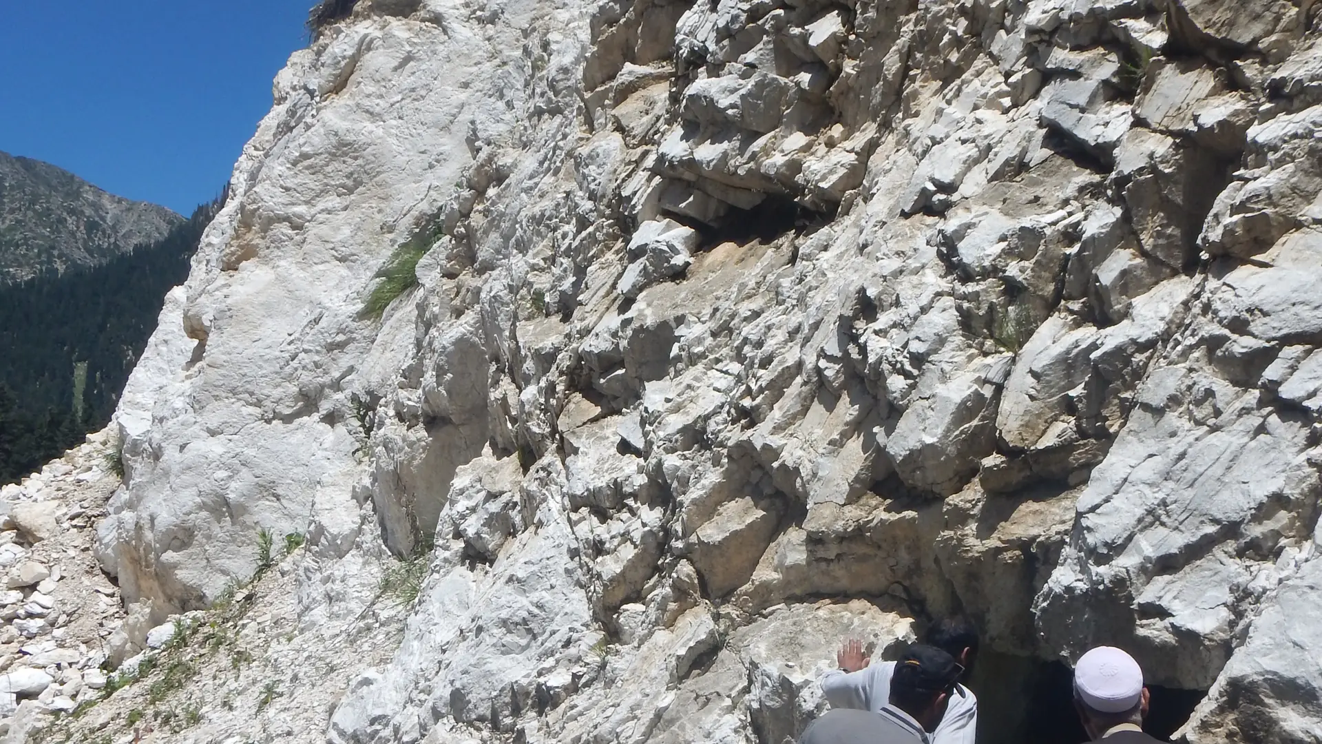 Men gathered at the entrance of an adit cut into a white rock cliff among pine trees.