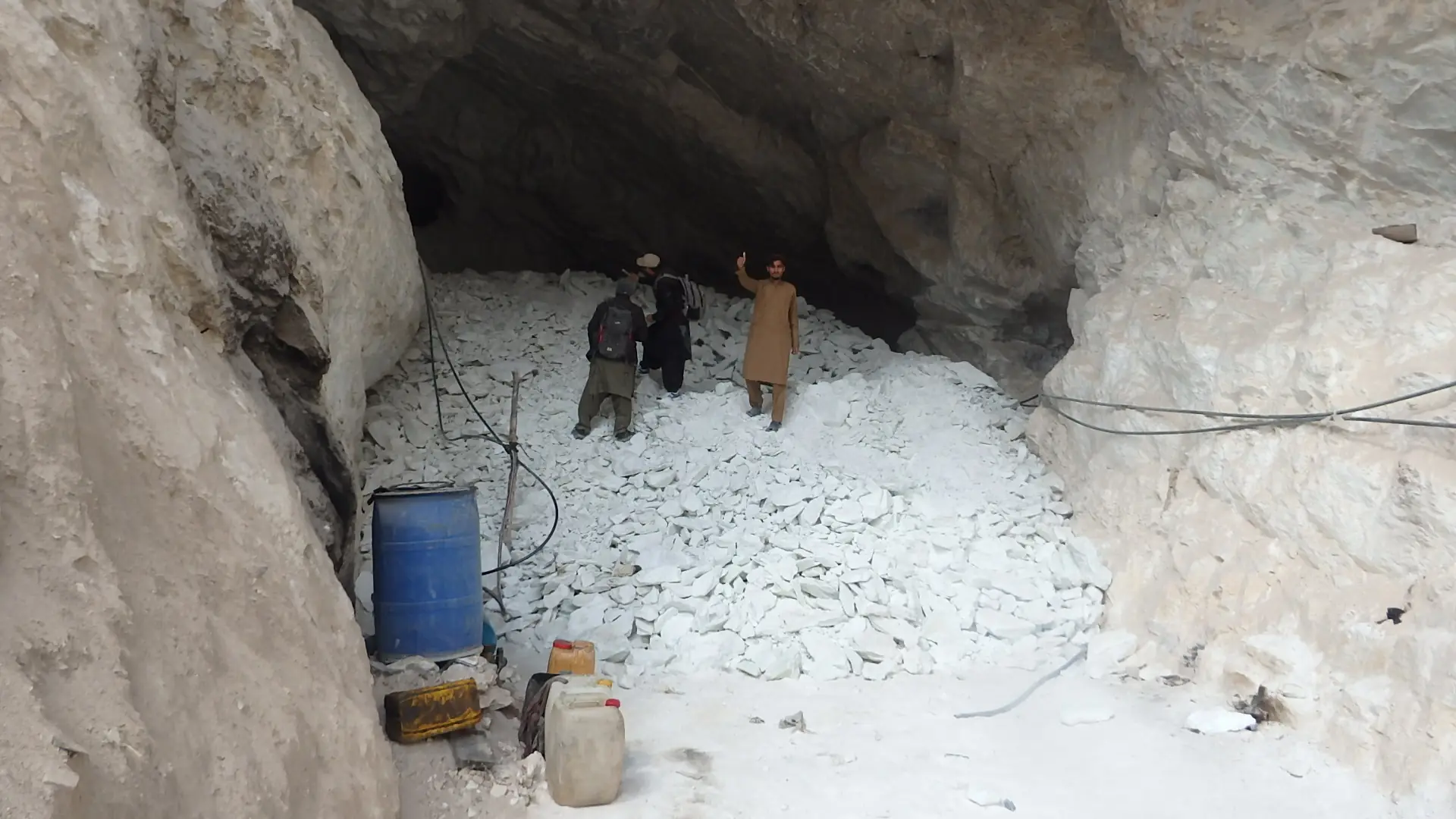 Two workers inside a large underground chamber floored with broken white rock, with a blue drum and hoses.