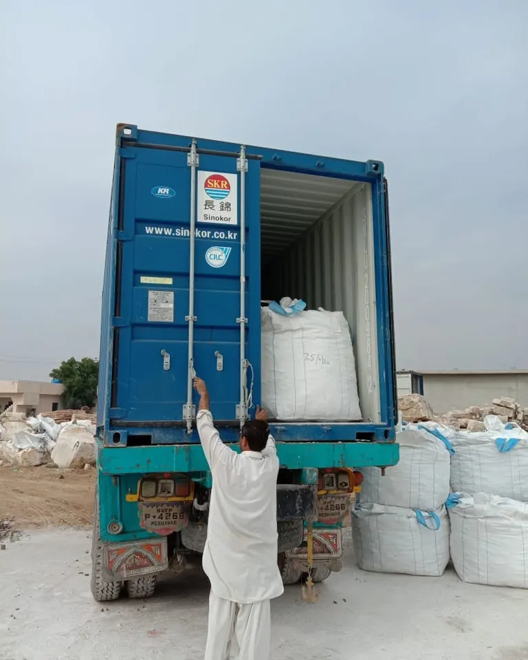 Worker loading white jumbo bags into a blue shipping container mounted on a truck.