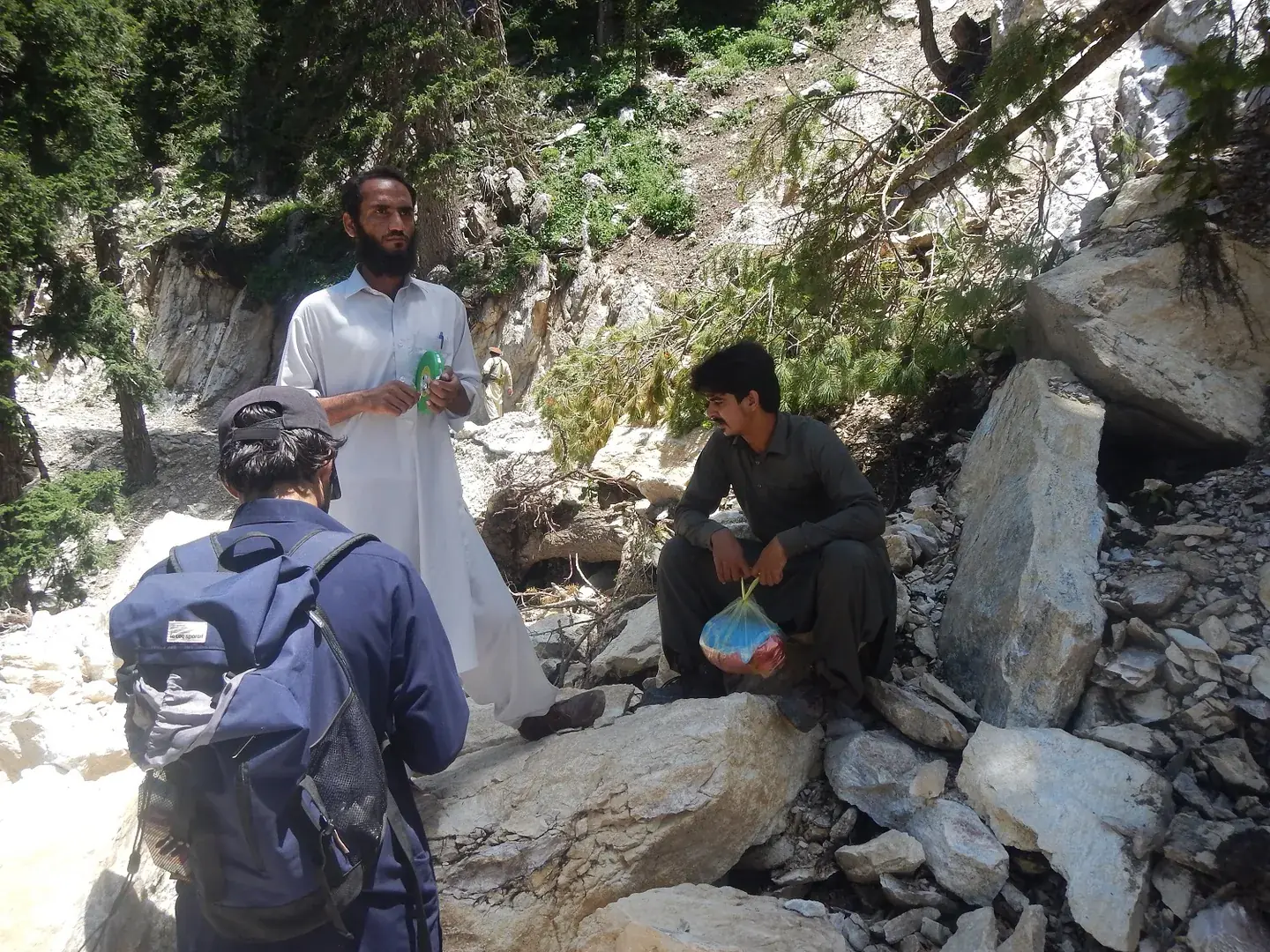 Field team with backpacks among boulders on a forested mountain slope.