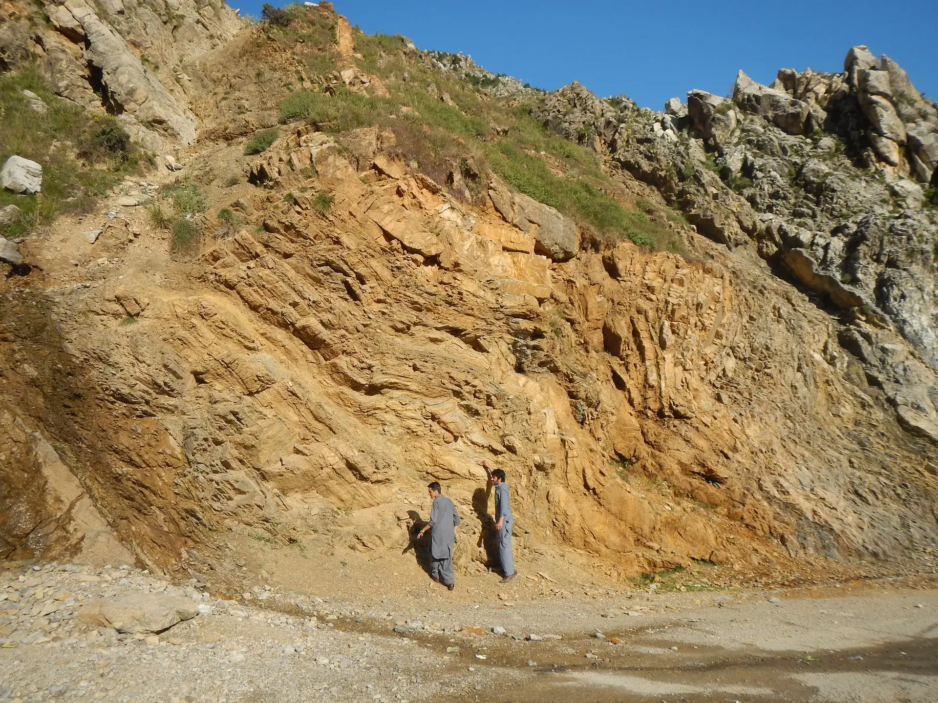 Two geologists at the base of a large folded orange-brown rock face.