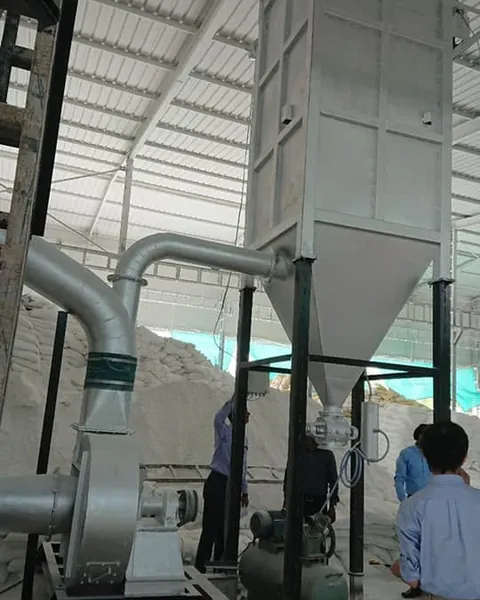 Grinding mill with cyclone and dust-collection hopper inside a processing shed, with two staff inspecting.