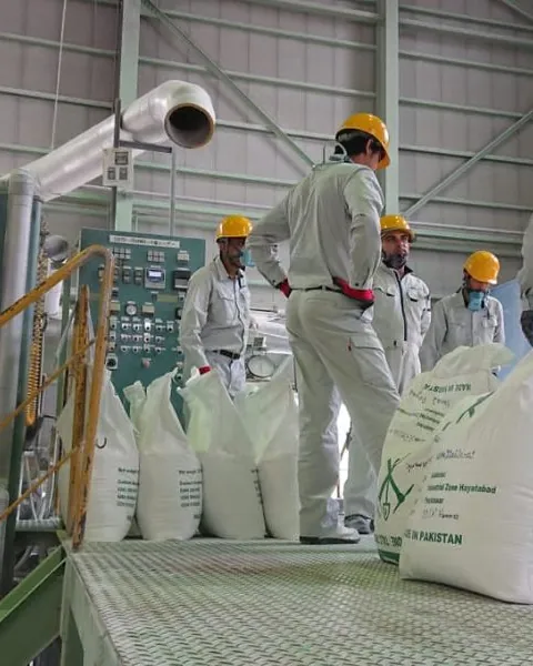 Workers in white uniforms and helmets filling and stacking SKZ talc powder bags in a large plant.